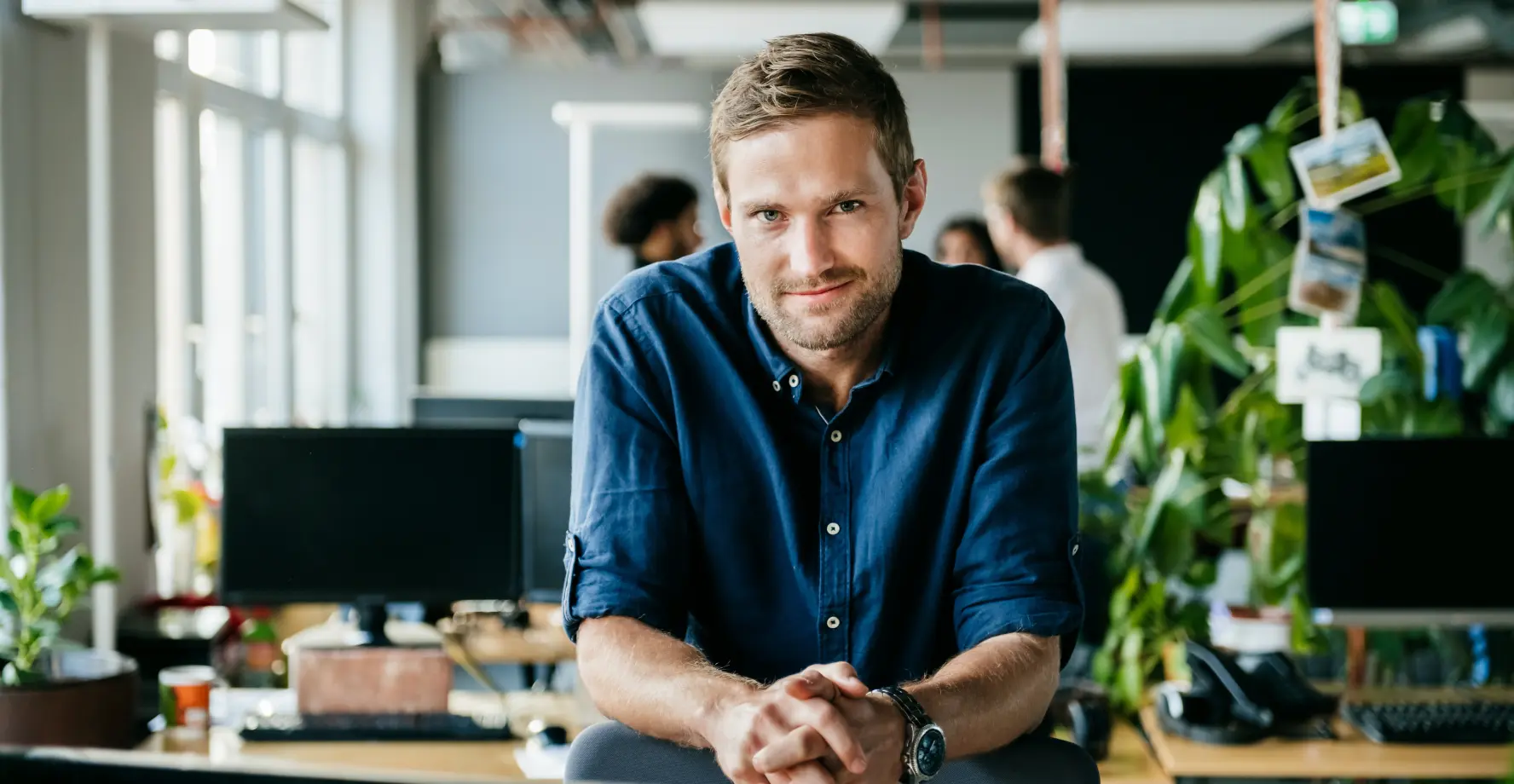 Mann ist mit Ellenbogen auf einen Schreibtisch gelehnt und blickt zufrieden in Richtung Kamera